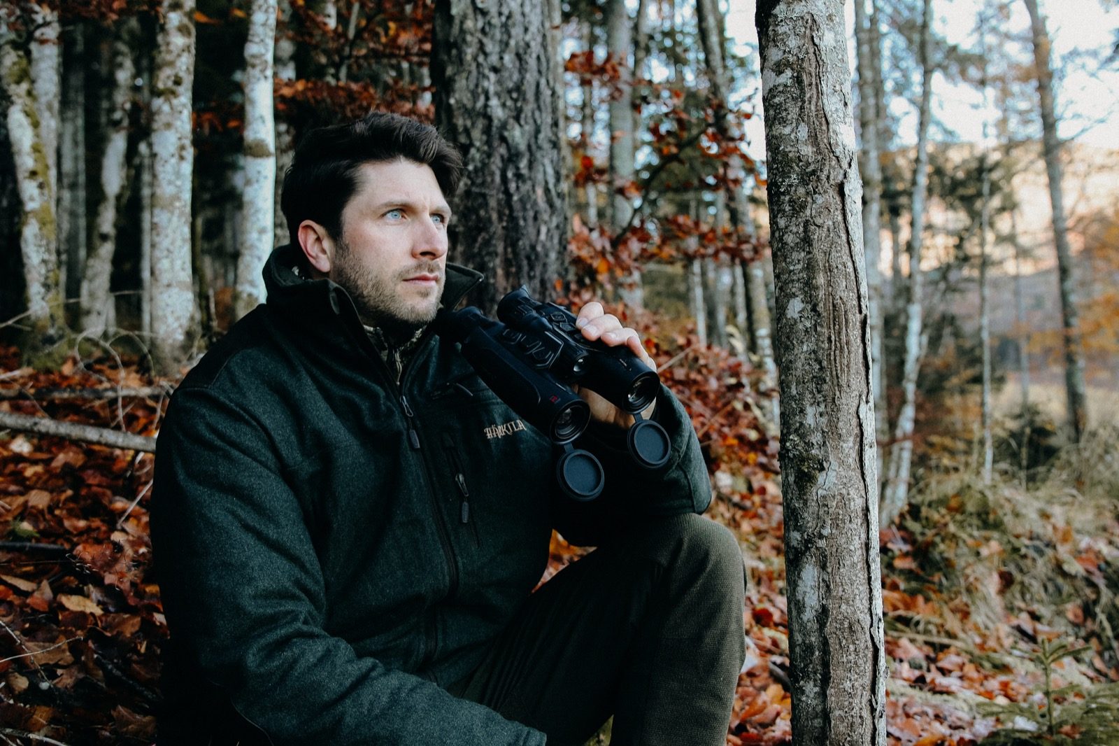 Jägare sitter lutad mot trädstam i höstskog och håller HIKMICRO Habrok 4K 2.0 HE25L